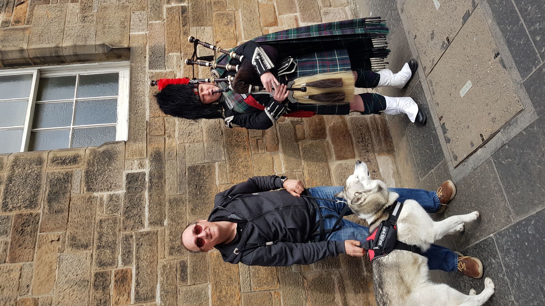 Scottish bagpipe player in traditional attire performing next to a tourist with a dog in Edinburgh.