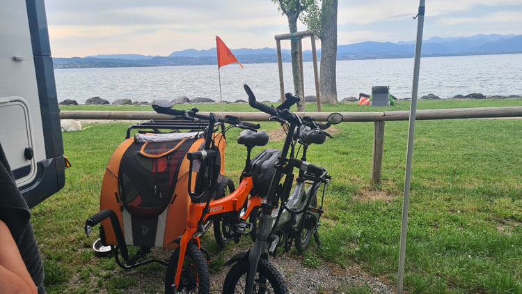 Two electric bikes with an orange cargo trailer parked on grass by a lake, near a paved area and a camper van in the foreground.
