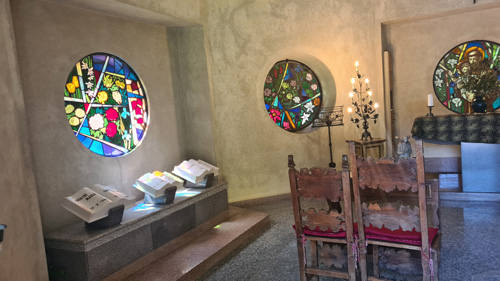 The image depicts a room featuring stained glass windows that likely allow colorful light to filter through. There are chairs arranged within the space, and various elements of interior design are present, including a coffee table and artwork on the walls. This indoor setting conveys an inviting atmosphere, reminiscent of a serene Giardini Botanici di villa Taranto chapel near Lake Maggiore.