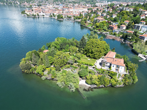 aerial shot of a lush, green island situated on a body of water, likely a lake. The island features a large villa or mansion with a terracotta roof, surrounded by meticulously landscaped gardens. The surrounding water is a deep teal, and the background shows a town or city nestled along the shoreline. The overall impression is one of tranquility, wealth, and idyllic natural beauty. Isolino di San Giovanni