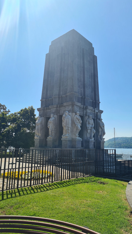 The image features a stone tower topped with statues, set against a backdrop of outdoor scenery. The sky is visible above, with clouds adding to the atmosphere. In the foreground, there are elements of grass and trees, along with other plants that enhance the natural setting. This monument serves as a significant landmark in the park near Lake Maggiore.
