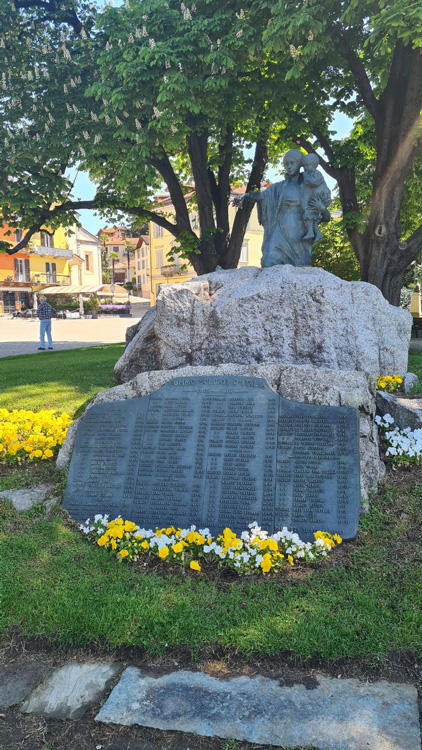 The image features a statue of a man and a child sitting on a rock, surrounded by natural elements. The setting includes trees, flowers, and grass, suggesting it is located in an outdoor park or cemetery near Lake Maggiore. This memorial sculpture serves as a tribute, enhancing the serene atmosphere of the area.