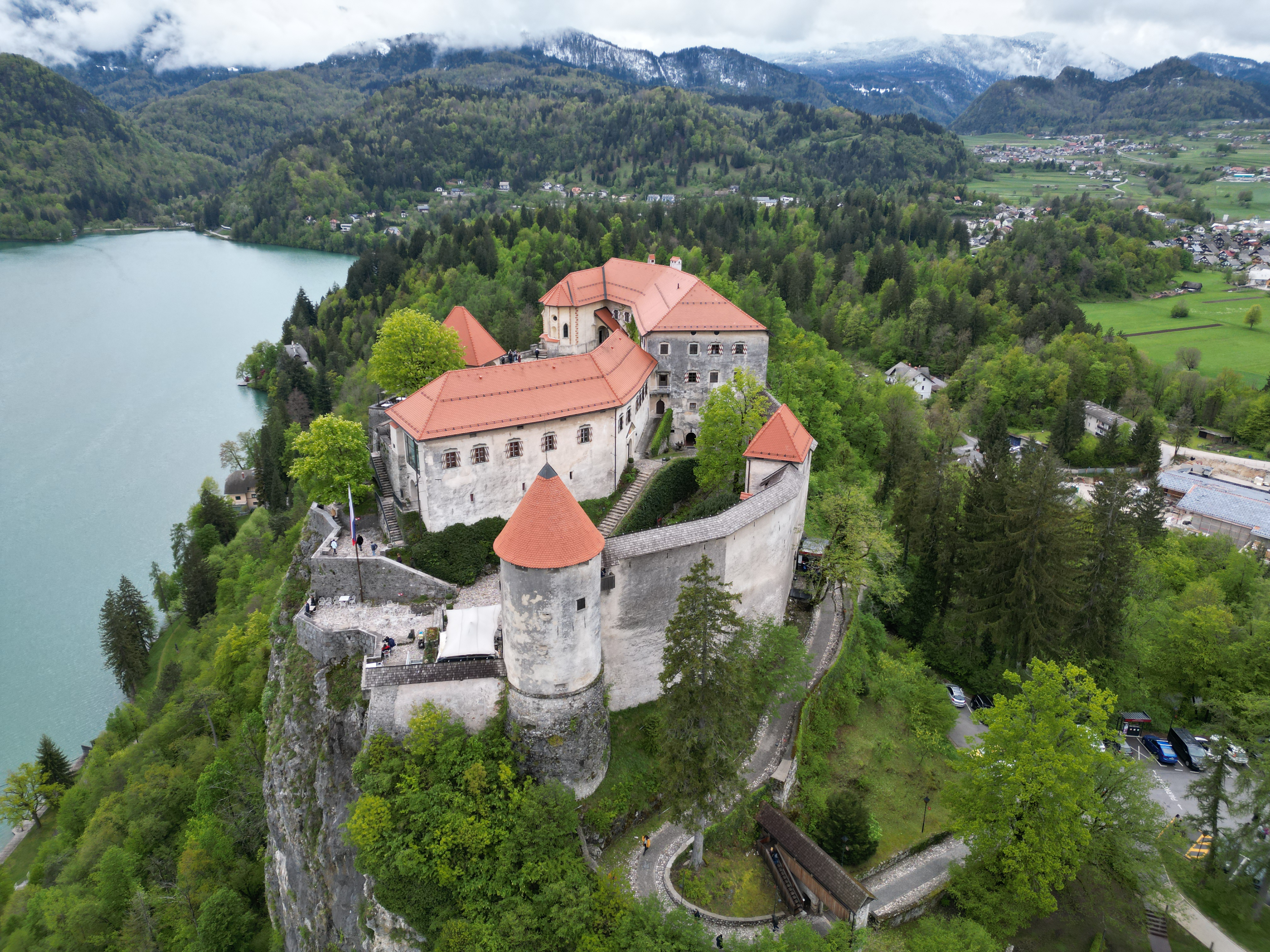 Aerial view of Bled Castle perched on a cliff, overlooking the turquoise waters of Lake Bled and surrounded by lush forests.