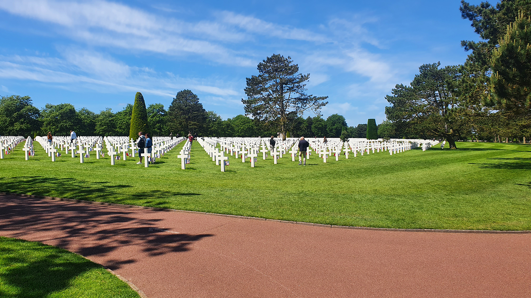 Rows of white crosses marking graves at the Normandy American Cemetery, with visitors walking among them, surrounded by green grass and trees under a clear blue sky.