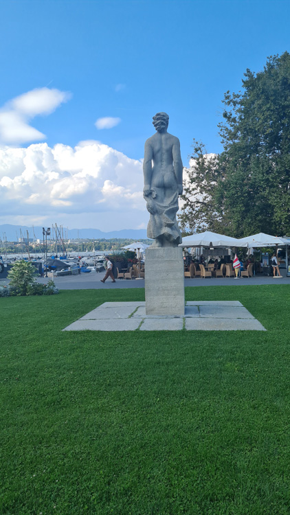 Statue in a park surrounded by open green space, trees, and clouds in the sky