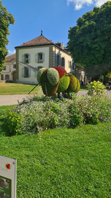 Large colorful bee sculpture in a vibrant garden with green grass, trees, shrubs, and a cloudy sky
