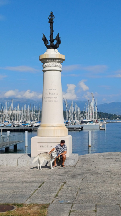 Person and dog seated on a bench in front of a statue at Port Noir, with lake and clear sky in the background