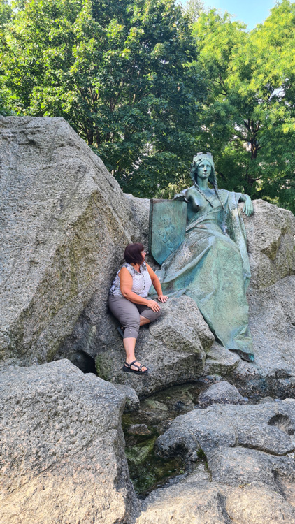 Lady posing beside the Universal Post Monument in Bern, surrounded by trees and rocky terrain on a hiking trail