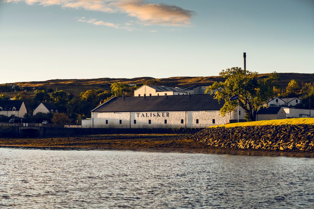 A scenic view of Talisker Distillery, located on the rugged shores of the Isle of Skye in Scotland, showcasing its iconic white buildings against a backdrop of rolling hills and a clear blue sky.