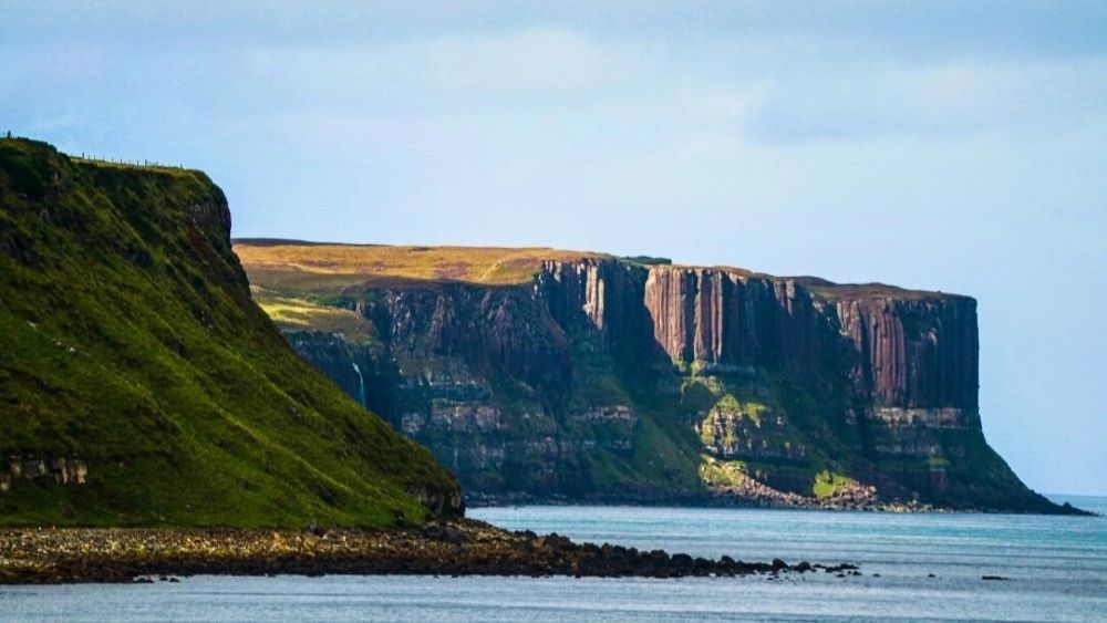 The dramatic landscape of the Trotternish Peninsula on the Isle of Skye, featuring rugged cliffs, rolling green hills, and the iconic rock formations of the Quiraing under a cloudy sky.